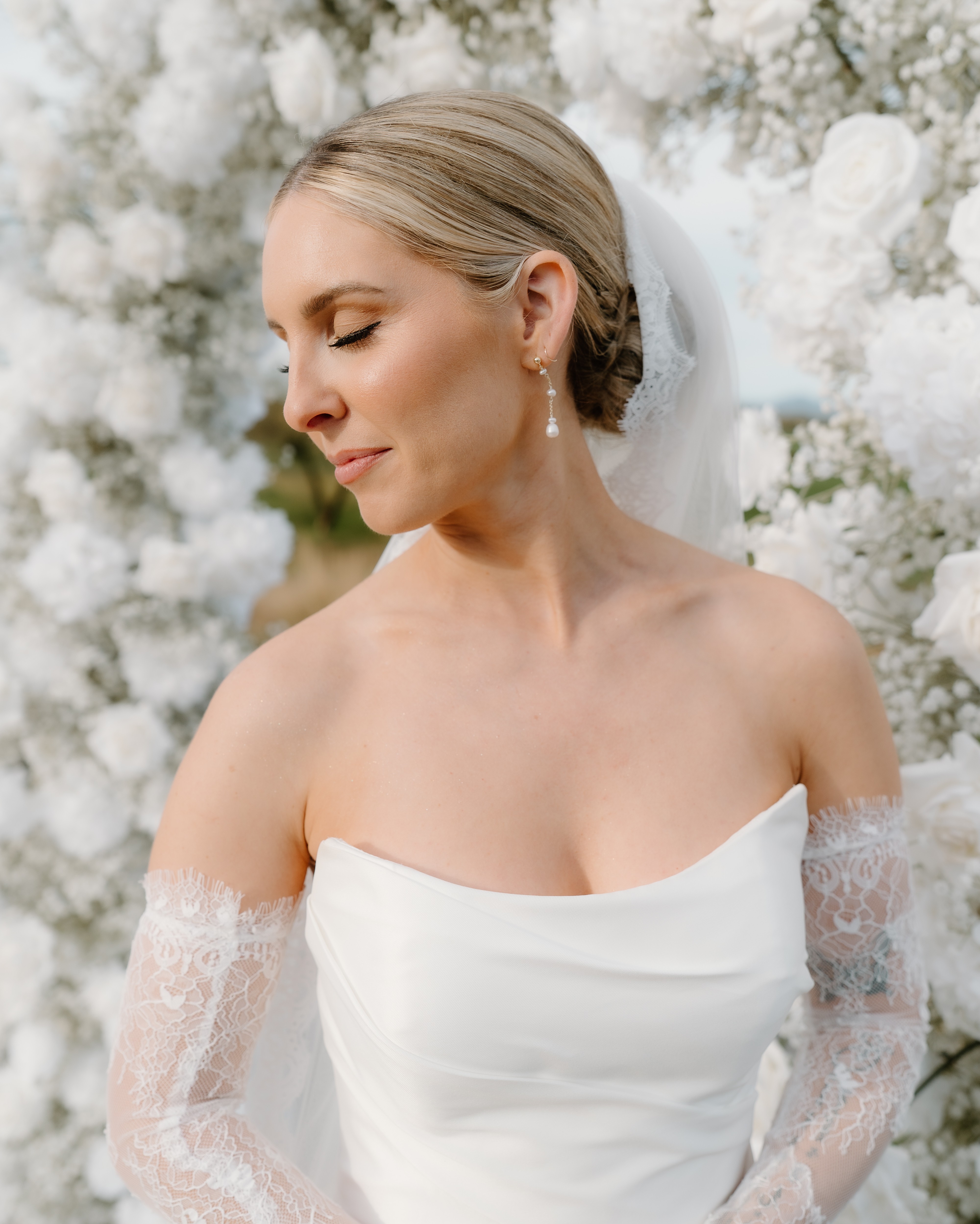 Close-up portrait of a bride with a sleek updo, soft makeup, and lace sleeves against white flowers.